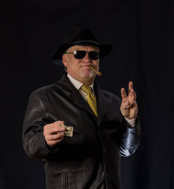 A man in black clothes, a black leather coat, hat. Ganster with a cigar. Mafiosi in black glasses on a dark background. Severe, strong, stylish, impressive, dangerous, unfriendly, evil, rude man - Photo, Image