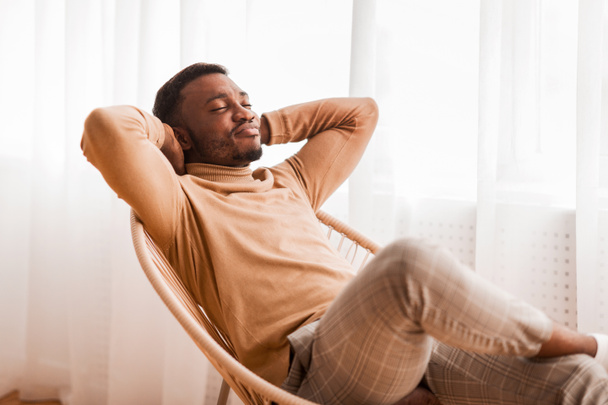 Черный парень расслабляется, сидя на современных волосах на фоне окон в помещении
 - Фото, изображение