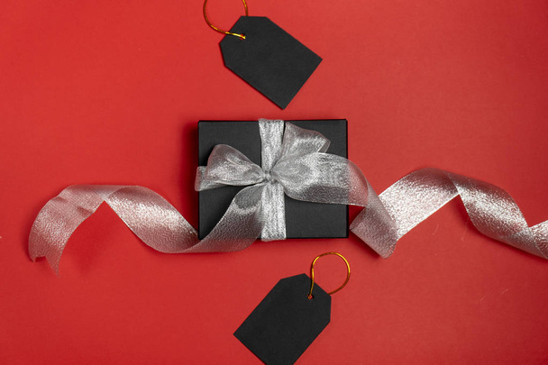 Gift box wrapped in black paper with silver ribbon and price tags on bright red background. Copy space and top view. Black friday box gift present isolated. - Fotoğraf, Görsel