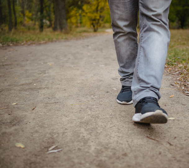 Ανδρικά πόδια σε τζιν και sneakers με τα πόδια έξω στην απόσταση στο έδαφος καλύπτονται με πεσμένα κίτρινα φύλλα. Φθινόπωρο έννοια άφιξης, κοντινό, αντίγραφο χώρου. - Φωτογραφία, εικόνα