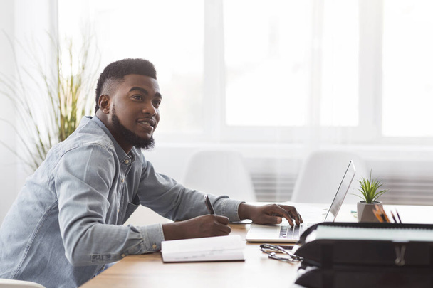 Musta mies tekee muistiinpanoja kannettavasta tietokoneesta työpaikalla
 - Valokuva, kuva