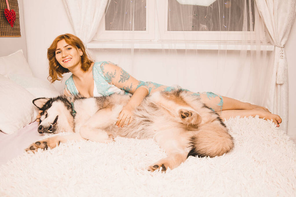 charming plus size girl with red hair in a nightgown posing with her large dog, a Malamute best friend in white bed in the bedroom - Photo, Image