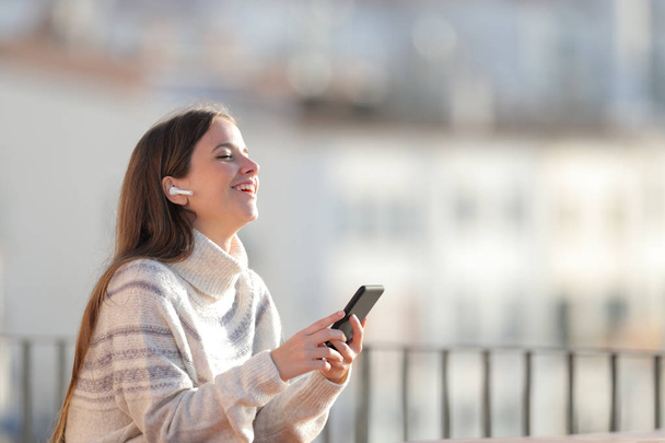 Happy woman listens to music with wireless earbuds in a balcony - Foto, Imagem
