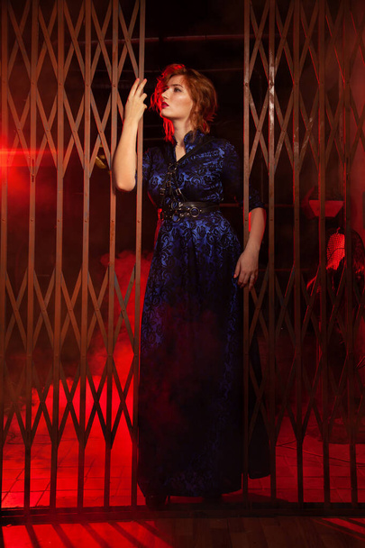 A vörös hajú plusz méretű nő estélyi hosszú ruha és bőr hámot áll közel a nagy kapuk egy fekete sikátorban éjszaka - Fotó, kép