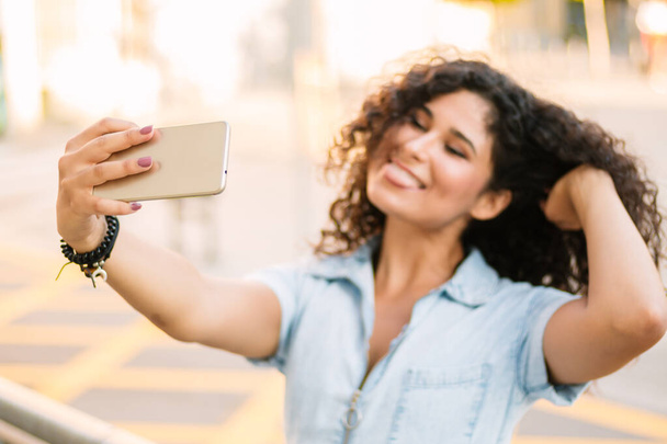 Ritratto di felice ragazza bruna riccia che si fa un selfie mentre mostra la lingua formica che si tocca i capelli per strada
 - Foto, immagini