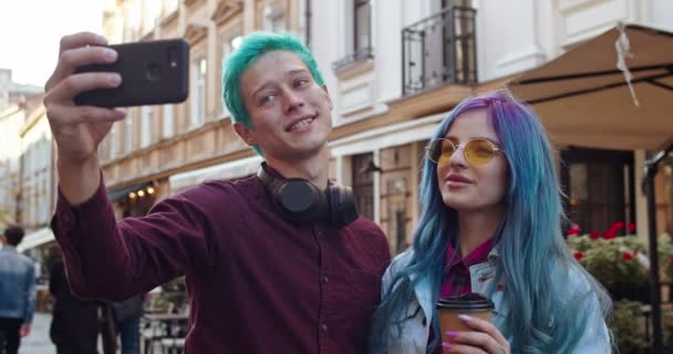 Close up of the young joyful Caucasian couple of hipsters with blue hair smiling and posing to the smartphone camera as taking selfie photos in the town. Outdoor. - Footage, Video