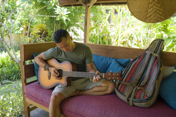 young handsome and happy mixed ethnicity man in hipster style chilling outdoors playing guitar relaxed at tranquil tropical garden enjoying holiday retreat  - Photo, image