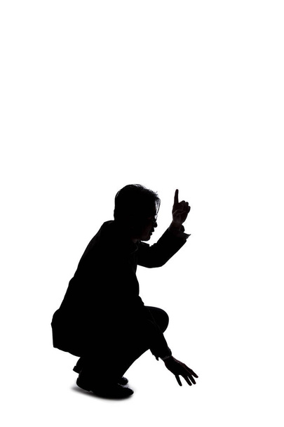 Silhouette of a backlit model posing as a businessman on a white background.  He is crouched on the ground like a detective looking or searching for something on the floor  - 写真・画像