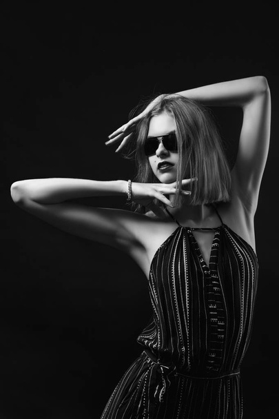 A young woman with black overalls and black glasses holds her hands near her head in the form of a frame and poses on a black isolated background. Fashion photography in black and white style. - Photo, Image