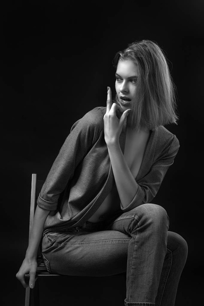 Sensuele Jonge vrouw in een grijze trui en jeans zit op een stoel en poseert op een zwarte geïsoleerde achtergrond. Fashion fotografie in zwart-wit stijl. - Foto, afbeelding