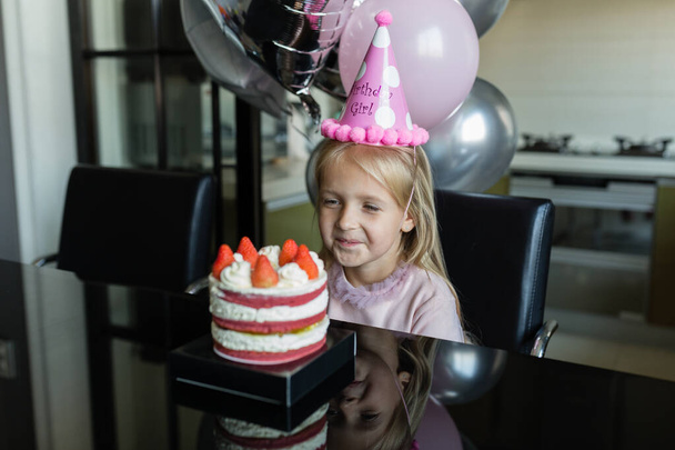Indoor shot of pretty joyful little girl with blonde hair blowing out the candle, celebrate 6 years old birthday, wear fashionable dress, have excited expressions. Happy childhood concept - Foto, Imagem