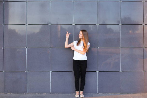 Молодая энергичная девушка подняла руки и указывает на рекламное пространство, стоящее именно в полном росте на фоне стены бизнес-центра
. - Фото, изображение