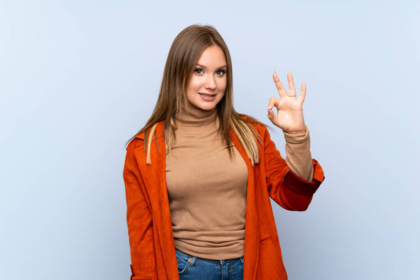 Teenager girl with coat over isolated blue background showing ok sign with fingers - Foto, Imagen