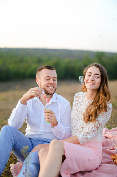 Young smiling man blowing soap bubbles near caucasian girl and sitting on plaid in steppe. - Foto, Imagem