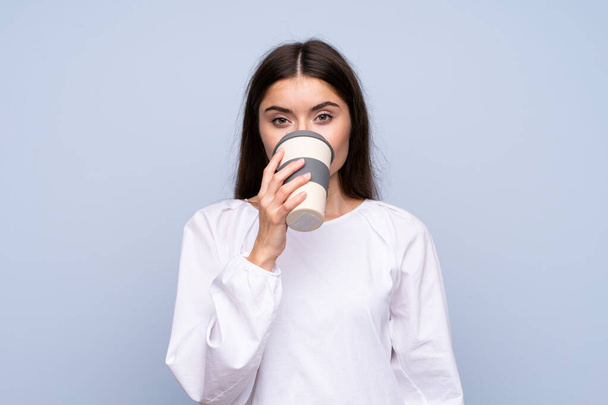Young woman over isolated blue background holding and drinking a coffee to take away - Foto, afbeelding