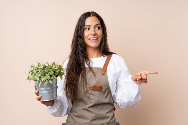 Mujer joven sosteniendo una planta sorprendida y señalando con el dedo hacia un lado
 - Foto, imagen