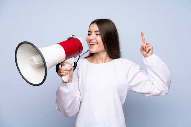 Young woman over isolated blue background shouting through a megaphone - Foto, Imagen