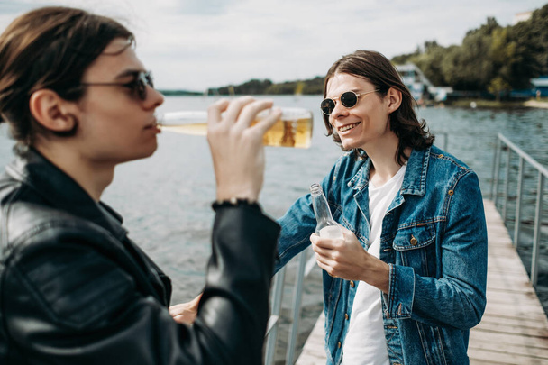 Twee stijlvolle tweeling broers hebben plezier drinken bier op de pier in de buurt van de rivier - Foto, afbeelding