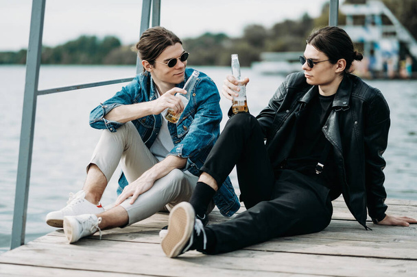 Twee stijlvolle tweeling broers hebben plezier drinken bier op de pier in de buurt van de rivier - Foto, afbeelding
