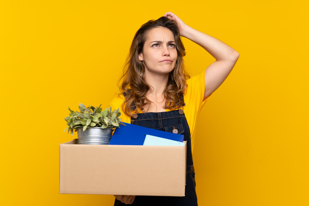 Young blonde girl making a move while picking up a box full of things having doubts and with confuse face expression - Photo, Image