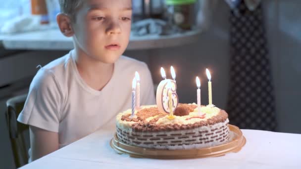 Little six-year-old caucasian boy in white t-shirt sitting at table at kitchen and blowing lighted candles on birthday cake, making wish. Candle in shape of number six on pie. Celebrating at home - Video, Çekim