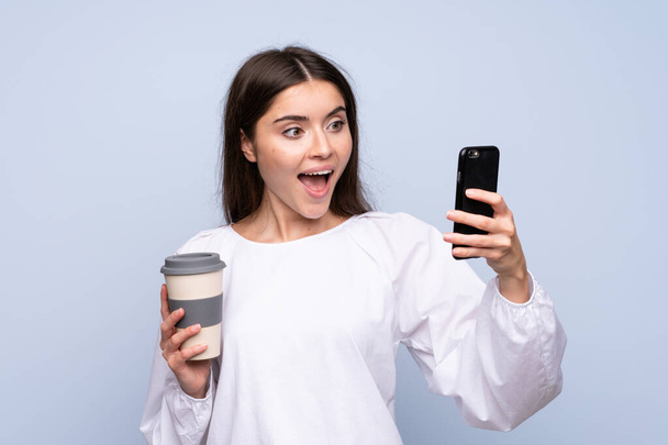 junge Frau über isoliertem blauem Hintergrund mit Kaffee zum Mitnehmen und einem Handy - Foto, Bild