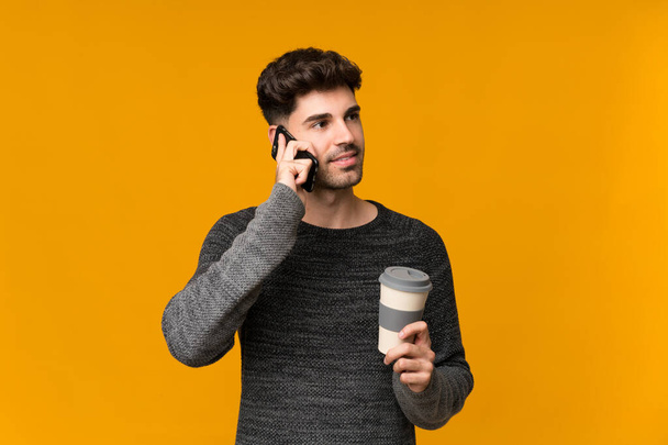Young man over isolated background holding coffee to take away and a mobile - Fotografie, Obrázek