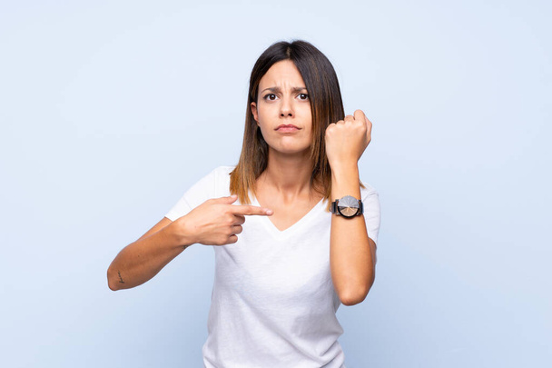 Young woman over isolated blue background showing the hand watch with serious expression serious because it is getting late - Foto, Imagem