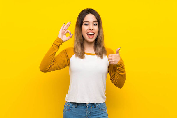 Csinos fiatal nő felett elszigetelt sárga fal mutató ok jel és hüvelykujj fel gesztus - Fotó, kép