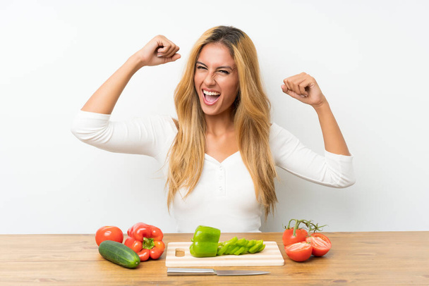 Young blonde woman with vegetables in a table celebrating a victory - Фото, зображення