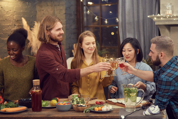 Multi-ethnic group of young people enjoying party together standing at table in modern interior and clinking glasses - Photo, Image