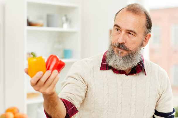 Portrait of bearded senior man holding fresh bell peppers while cooking vegetables in kitchen, copy space - Фото, зображення