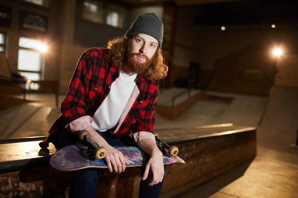 Портрет современного бородатого человека, смотрящего в камеру, сидящего на рампе в тусклом скейт-парке, копировальное пространство
 - Фото, изображение