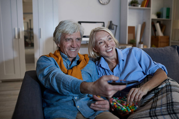 Retrato de una pareja de ancianos sonrientes viendo la televisión juntos sentados en un cómodo sofá en la oscuridad, espacio para copiar
 - Foto, imagen