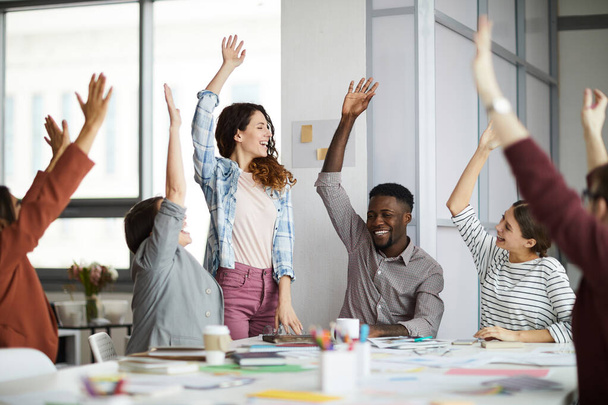 Портрет творческой команды предпринимателей, поднимающей руки при праздновании успеха за столом встреч в современном офисе, акцент на улыбающейся молодой женщине, стоящей в центре
 - Фото, изображение