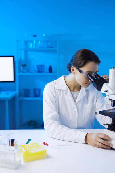 Porträt einer jungen Wissenschaftlerin, die unter dem Mikroskop forscht, während sie im von blauem Licht beleuchteten medizinischen Labor forscht, Kopierraum - Foto, Bild