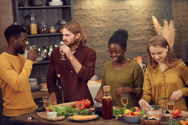 Grupo multiétnico de jóvenes que cocinan la cena juntos de pie en la mesa en el interior moderno y charlando
 - Foto, Imagen
