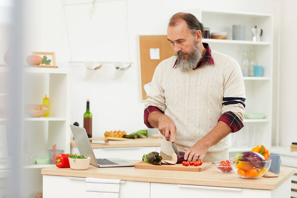 Waist up portrait of bearded senior man watching video recipe via laptop while cooking healthy salad in kitchen, copy space - Fotografie, Obrázek