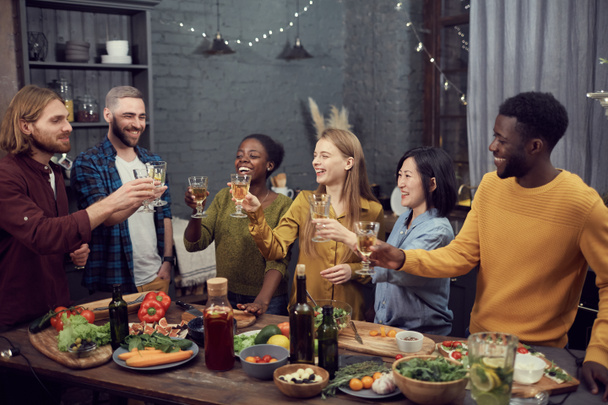 Grupo multi-étnico de jovens sorridentes que gostam de jantar juntos em pé à mesa no interior moderno e brindar com copos de vinho, espaço de cópia
 - Foto, Imagem