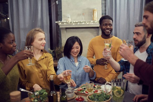 Grupo multiétnico de jóvenes disfrutando de la cena juntos de pie en la mesa en el interior moderno y sosteniendo copas de vino
 - Foto, Imagen