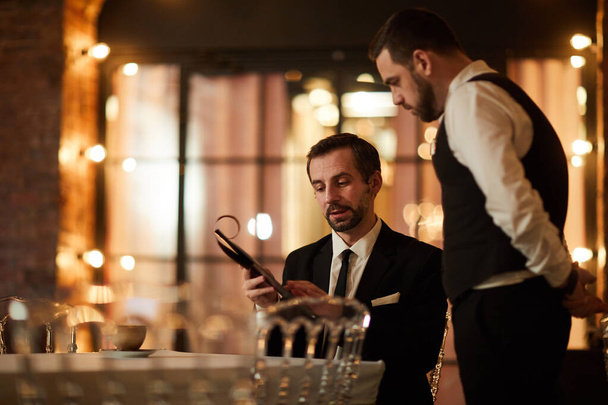 Portret van volwassen bebaarde zakenman op zoek naar menu tijdens het bestellen van diner in luxe restaurant, kopieer ruimte - Foto, afbeelding