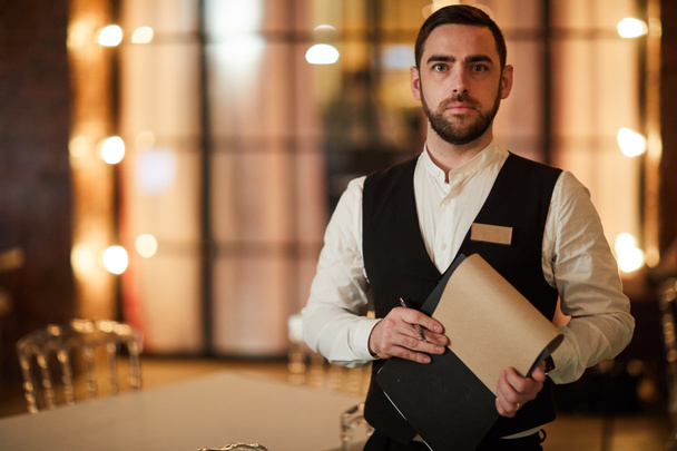 Waist up portrait of handsome waiter looking at camera with serious face expression and holding clipboard while posing in luxury restaurant, copy space - Foto, Imagem