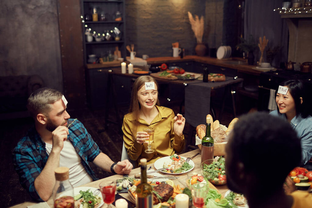 Grupo multiétnico de jóvenes que juegan a adivinar mientras están sentados en la mesa durante la cena en el cuarto oscuro, espacio para copiar
 - Foto, imagen