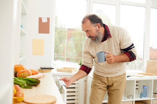 Retrato de vista lateral de hombre mayor barbudo abriendo armarios de cocina mientras busca desayuno por la mañana, espacio para copiar
 - Foto, Imagen