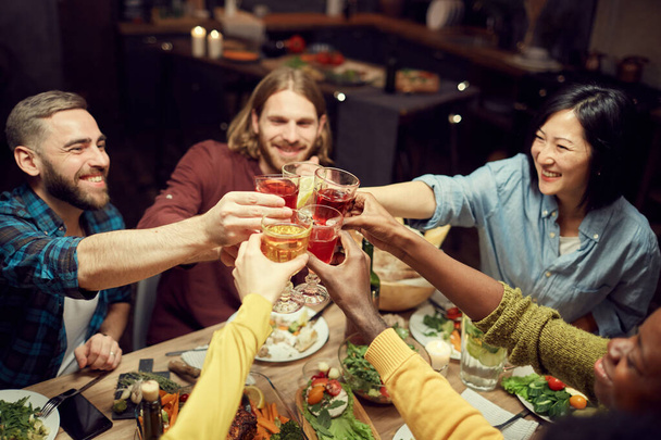 Korkea kulma näkymä monikansallinen ryhmä ihmisiä nostaa lasit ja paahtoleipää juhla istuu ruokapöydässä hämärässä huoneessa, kopioi tilaa
 - Valokuva, kuva