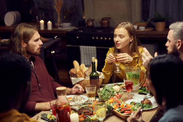 Groupe de jeunes gens modernes profitant du dîner et du vin ensemble assis à table dans une pièce faiblement éclairée, espace de copie
 - Photo, image