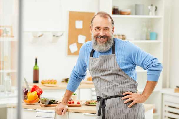 Retrato de la cintura hacia arriba del hombre mayor barbudo que usa delantal mirando alegremente a la cámara mientras cocina en casa, espacio para copiar
 - Foto, imagen