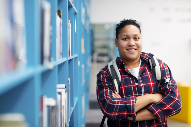 Waist up portrait of mixed-race college student wearing backpack and smiling at camera cheerfully while posing in library, copy space - 写真・画像