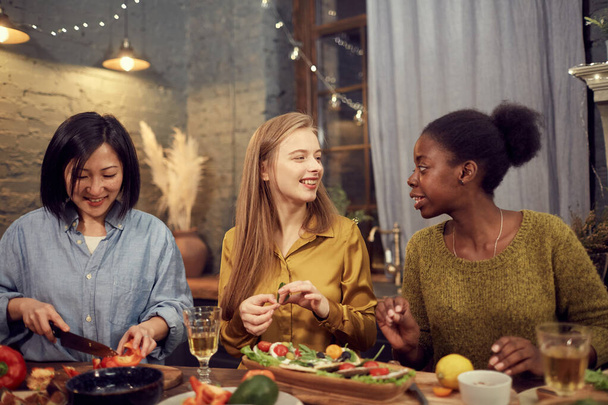 Portret van drie jonge vrouwen koken diner voor huisfeest terwijl ze in de rij staan aan tafel en vrolijk chatten - Foto, afbeelding