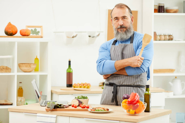 Taillenhoch Porträt eines bärtigen älteren Mannes, der zu Hause kocht und in die Kamera schaut, während er selbstbewusst in der Küche posiert, Kopierraum - Foto, Bild